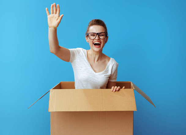 happy young woman in white t-shirt get out of a cardboard box and against blue background - Photo, Image