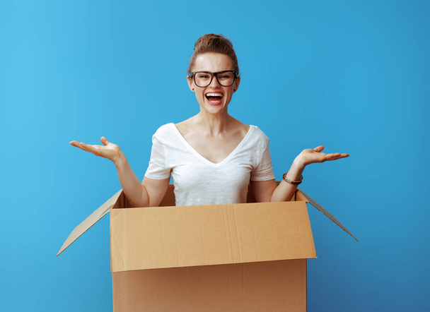 happy modern woman in white t-shirt presenting something on empty palm pops out of a cardboard box and against blue background - Photo, Image