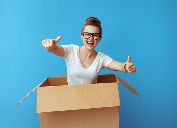 smiling modern woman in white t-shirt pops out of a cardboard box and showing thumbs up against blue background - Zdjęcie, obraz