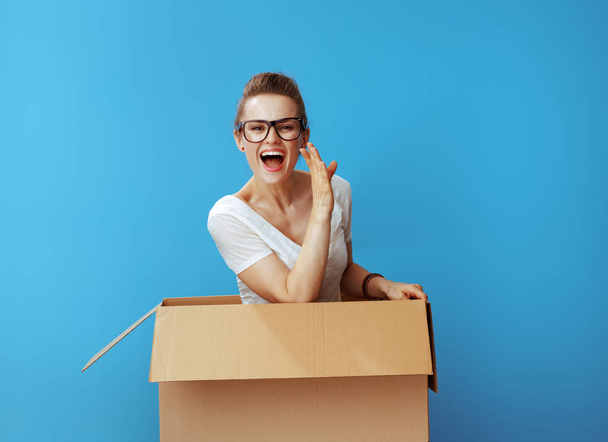 happy modern woman in white t-shirt in a cardboard box telling exciting news against blue background - Foto, immagini
