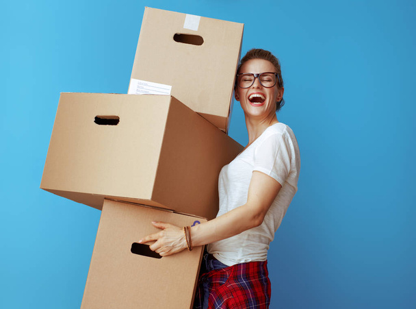 ευτυχισμένος νεαρή γυναίκα στο λευκό t-shirt εκμετάλλευση σωρό κουτιά από χαρτόνι που απομονώνονται σε μπλε - Φωτογραφία, εικόνα