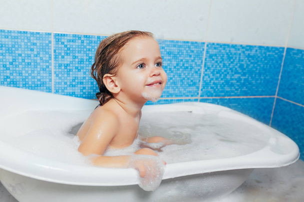 Fun cheerful happy toddler baby taking a bath playing with foam bubbles. Little child in a bathtub. Smiling kid in bathroom on blue background. Hygiene and health care - Photo, Image