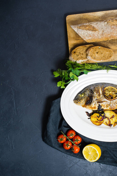 Печёная рыба Дорадо на белой тарелке, гарнир из жареной картошки. Темный фон, вид сверху
 - Фото, изображение