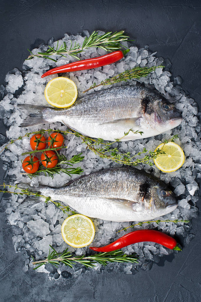 Dorado pescado crudo sobre hielo, ingrediente romero, limón, tomates, chile. Fondo oscuro, vista superior
 - Foto, Imagen