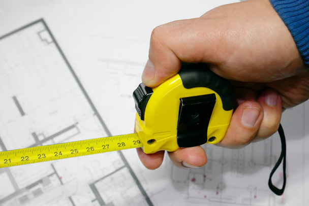 Рука будівельника тримає вимірювальну стрічку над планом будівництва і підлоги на задньому плані
.  - Фото, зображення
