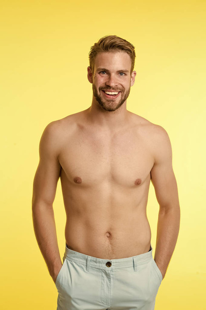 Man on smiling face puts hands in pockets, yellow background. Guy with bristle on face and smooth skin on torso posing in pants. Hair removal concept. Man looks attractive with smooth sexy torso - Foto, Imagem