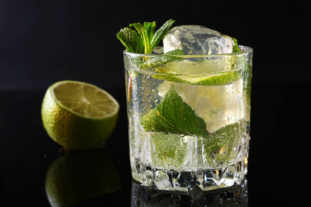 Glass of cocktail with rum, lime, ice cubes and mint leaves on black mirror background. - Foto, afbeelding