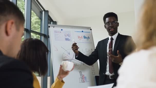 Young afro-american manager of company presenting new plan for project for team explaining business strategy using flipchart in office. Handsome speaker with flip chart reporting during the conference - Footage, Video