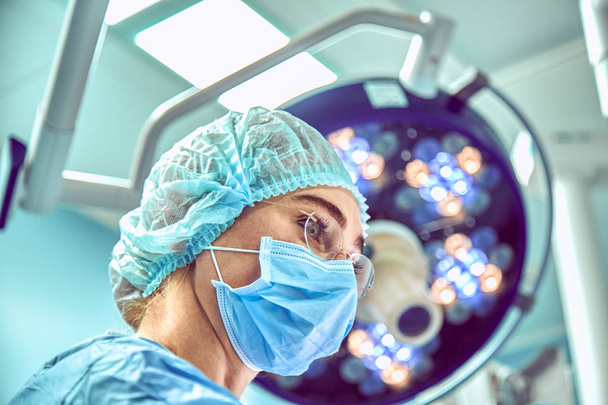 Close up portrait of young female surgeon doctor wearing protective mask and hat during the operation. Healthcare, medical education, surgery concept - Фото, изображение