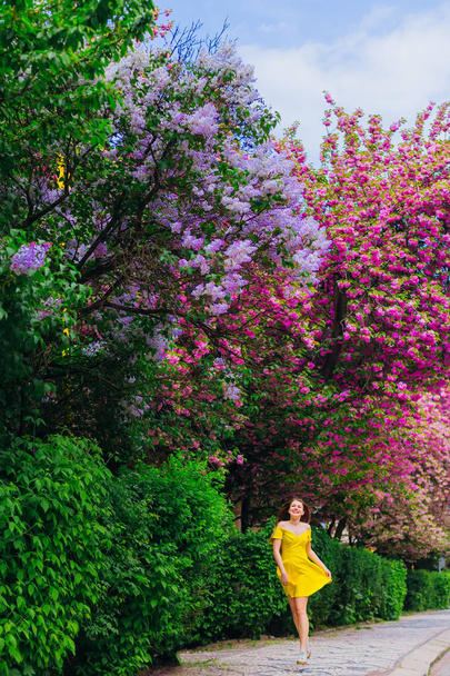 das Mädchen in einem stylischen Kleid läuft den Bürgersteig entlang und blickt in die Nähe der Bäume mit Blumen - Foto, Bild