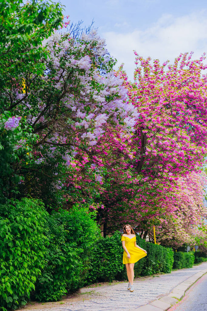 The girl in a stylish dress runs along the sidewalk and looks up near the trees with flowers - Photo, Image