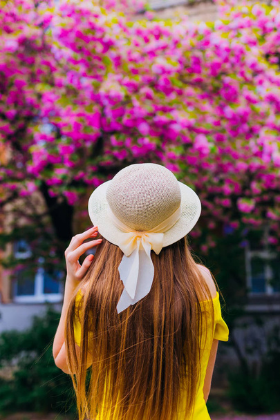 Close-up of a girl in a yellow dress and hat standing back to the camera and holding her hand on a hat on the background of a building and Japanese cherry - Photo, image