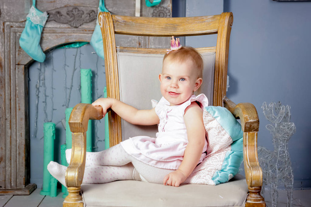 Portrait of a cute little european blond princess girl with a crown  in a beautiful dress sitting on a vintage chair in a studio decorated in the Christmas theme with toys, christmas tree and fireplace - Foto, afbeelding