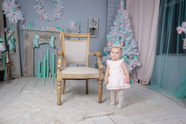 Portrait of a cute little european blond princess girl with a crown  in a beautiful dress sitting on a vintage chair in a studio decorated in the Christmas theme with toys, christmas tree and fireplace - Foto, immagini