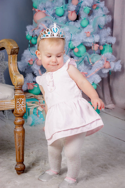 Portrait of a cute little european blond princess girl with a crown  in a beautiful dress sitting on a vintage chair in a studio decorated in the Christmas theme with toys, christmas tree and fireplace - Photo, Image