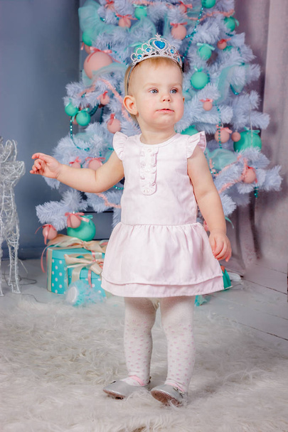 Portrait of a cute little european blond princess girl with a crown  in a beautiful dress in a studio decorated in the Christmas theme with toys and christmas tree - Photo, Image
