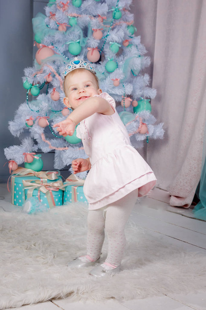 Portrait of a cute little european blond princess girl with a crown  in a beautiful dress in a studio decorated in the Christmas theme with toys and christmas tree - Photo, Image
