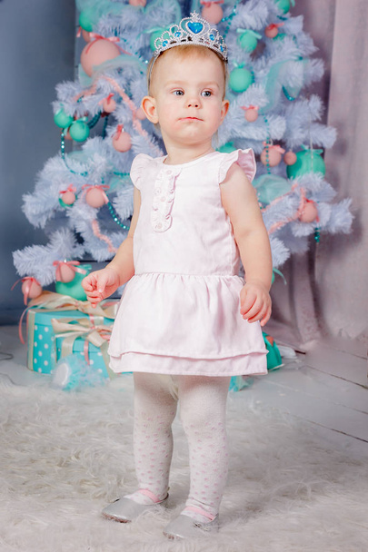 Portrait of a cute little european blond princess girl with a crown  in a beautiful dress in a studio decorated in the Christmas theme with toys and christmas tree - Фото, изображение