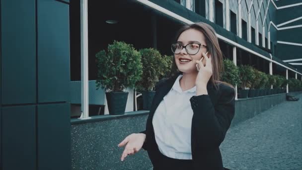 Young pretty smart business girl walking in business clothes. During this she is talking on the phone. - Video