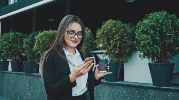 Young pretty smart business girl walking in business clothes. During this she keeps coffee and looks into the phone. - Video