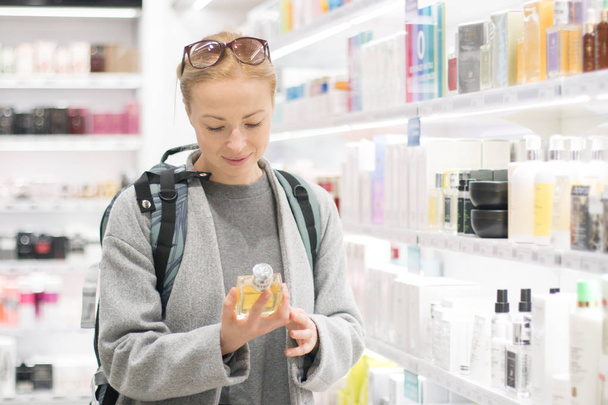 Blond young female traveler wearing travel backpack choosing perfume in airport duty free store. - Foto, imagen