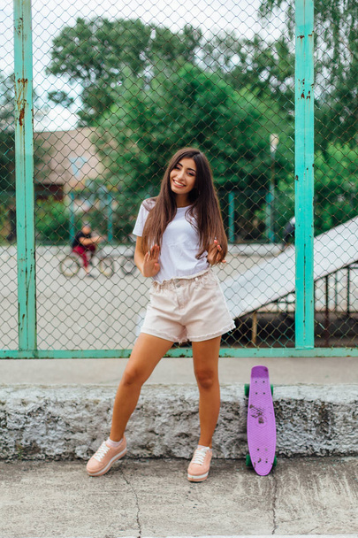 Summer lifestyle image of trendy pretty young girl standing next to the scateboard coart with her purple plastic scateboard. - Foto, imagen