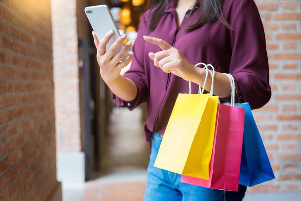 Nő pillantást mozgatható telefon-val paperbags a plázában, miközben élvezi a nap bevásárló - Fotó, kép