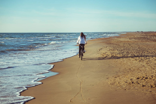 Woman rides a Bicycle along the sea on a sandy beach. Back view. - 写真・画像
