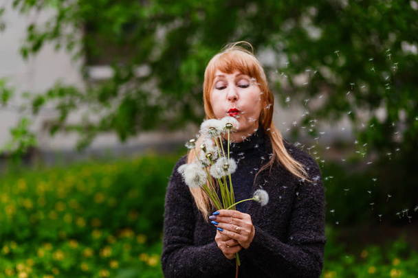middle-aged woman blowing off dandelion seeds outdoors blurred background - Foto, Imagen