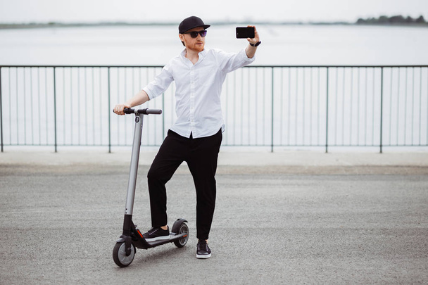Modern man in stylish outfit making selfie while standing at the street with electric scooter - Foto, immagini