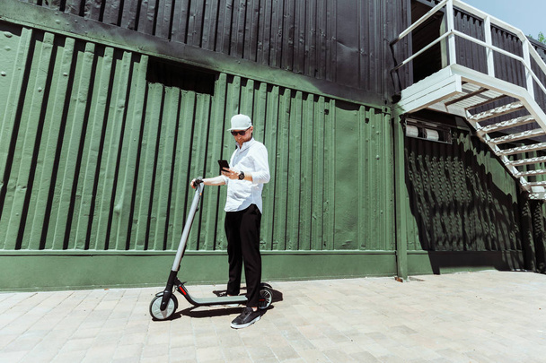 Modern man in stylish outfit using his smartphone while standing at street with electric scooter - Fotó, kép