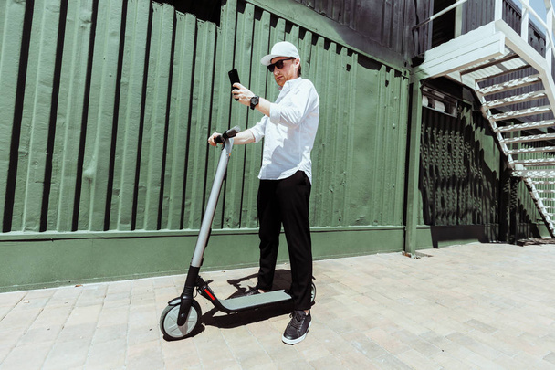 Modern man in stylish outfit using his smartphone while standing at street with electric scooter - Foto, Imagem