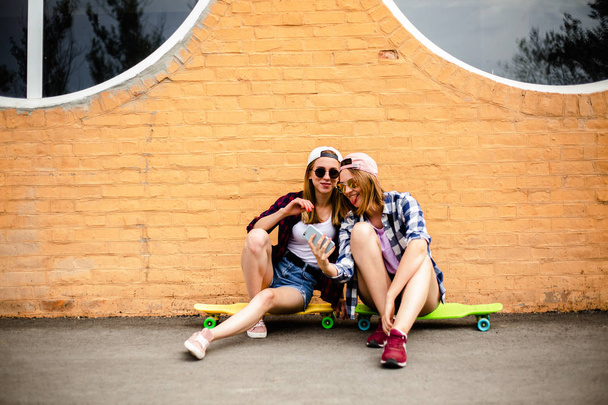 Two young happy girl friends in hipster outfit sitting on longboards and making selfie on phone - Photo, Image