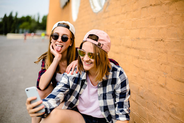 Two young happy girl friends in hipster outfit sitting on longboards and making selfie on phone - Zdjęcie, obraz