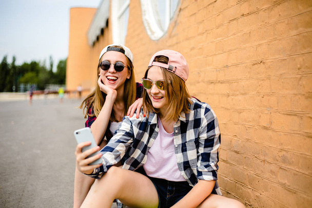 Two young happy girl friends in hipster outfit sitting on longboards and making selfie on phone - Foto, immagini