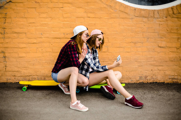 Two young happy girl friends in hipster outfit sitting on longboards and making selfie on phone - Фото, изображение
