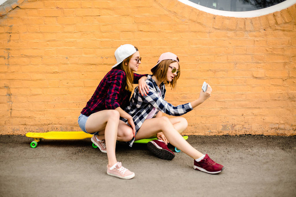 Two young happy girl friends in hipster outfit sitting on longboards and making selfie on phone - Foto, imagen