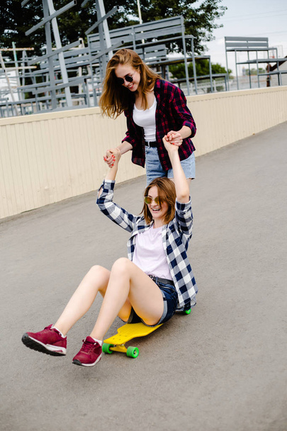 Two smiling female friends having fun riding yellow longboard on the street. Friendship concept - Zdjęcie, obraz