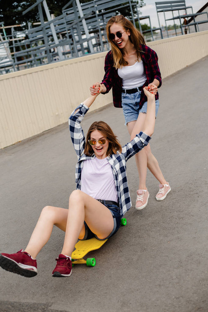 Two smiling female friends having fun riding yellow longboard on the street. Friendship concept - Photo, image