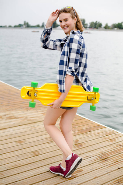 Young cheerful girl in hipster outfit holding yellow longboard in his hand and walking on a wooden pier - Photo, Image