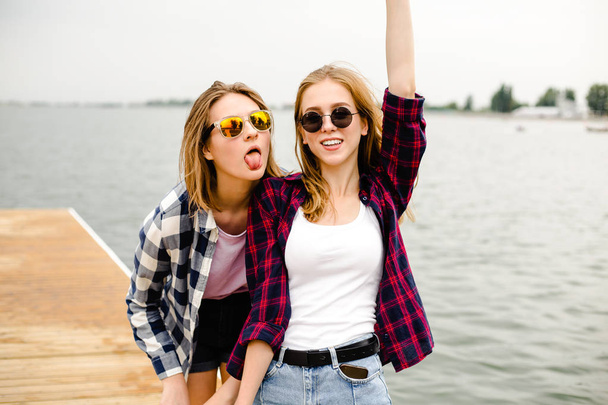 Two cheerful happy skater girls in hipster outfit having fun on a wooden pier during summer vacation - 写真・画像