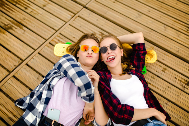Two stylish young urban girls with longboards lie on the wooden flooring in the street. Friends have fun and spend time together in the summer during the holidays - Foto, imagen