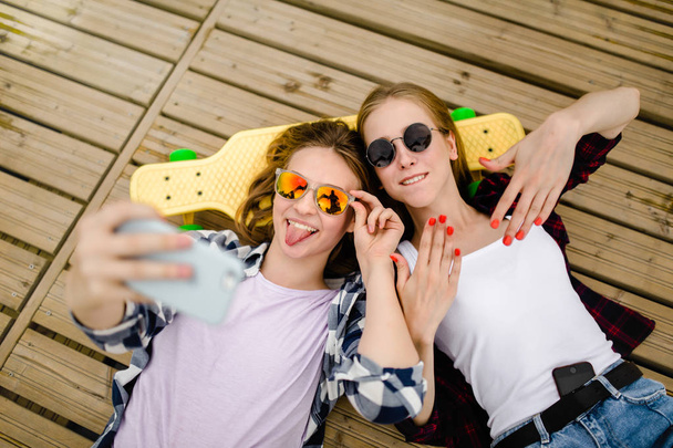 Two young girl in hipster outfit making selfie while lying with on wooden pier - Foto, imagen