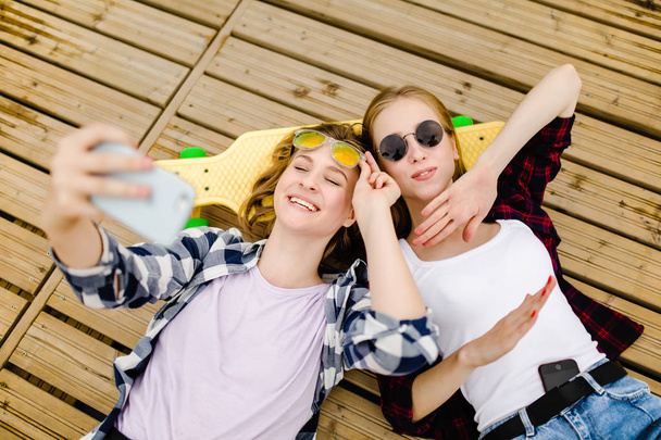 Two young girl in hipster outfit making selfie while lying with on wooden pier - Fotografie, Obrázek