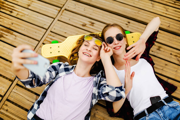 Two young girl in hipster outfit making selfie while lying with on wooden pier - Zdjęcie, obraz
