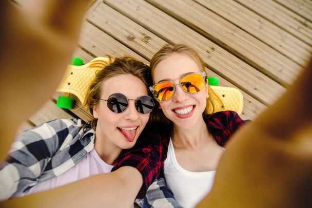 Two young girl in hipster outfit making selfie while lying with on wooden pier - Foto, Imagen