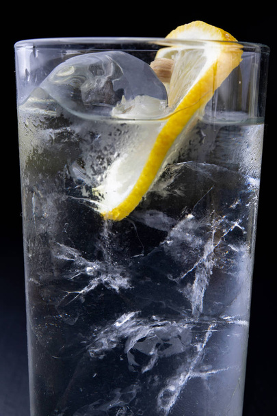 Cold drink in a glass on the kitchen table. Lemon and ice with water the best way to cool off on a summer day. Dark background. - Foto, Imagen