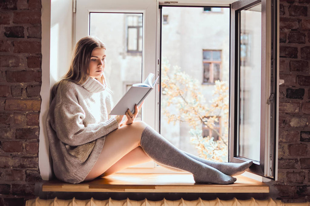 Ładna dziewczyna w ciepły sweter i skarpetki czyta książkę, siedząc na parapecie przy otwartym oknie - Zdjęcie, obraz