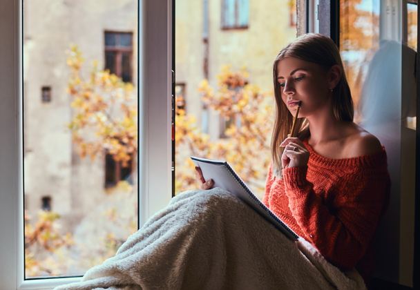 Menina em uma camisola quente cobriu as pernas com um cobertor, faz anotações em seu caderno sentado no peitoril da janela ao lado da janela aberta
 - Foto, Imagem