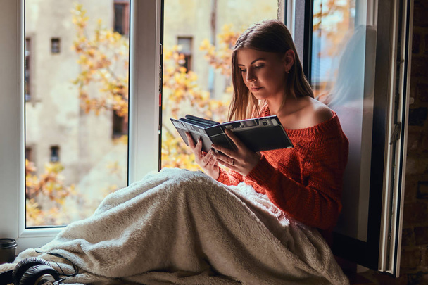 Νεαρή γυναίκα σε ένα ζεστό πουλόβερ που καλύπτονται στα πόδια της με μια κουβέρτα, βλέπουν το άλμπουμ φωτογραφιών, κάθεται στο περβάζι - Φωτογραφία, εικόνα